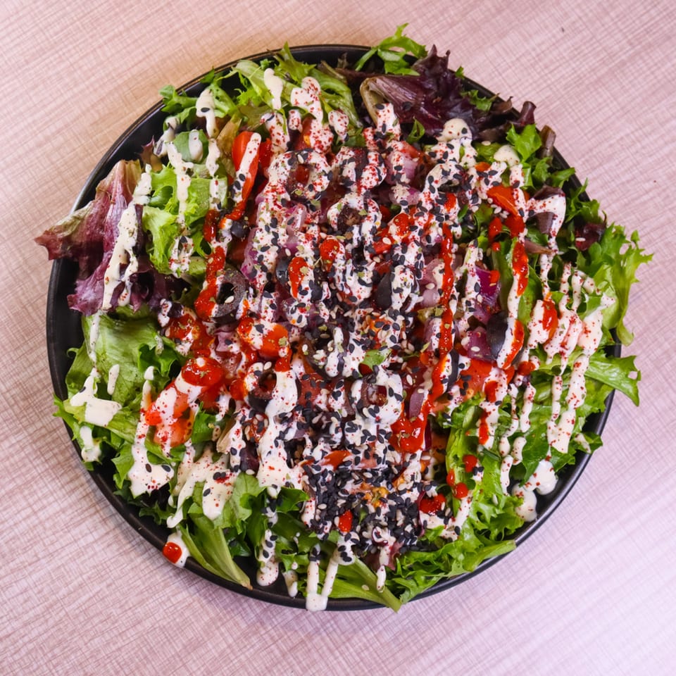 A vibrant salad with mixed greens, red sauce, white dressing, and black sesame seeds in a black bowl.