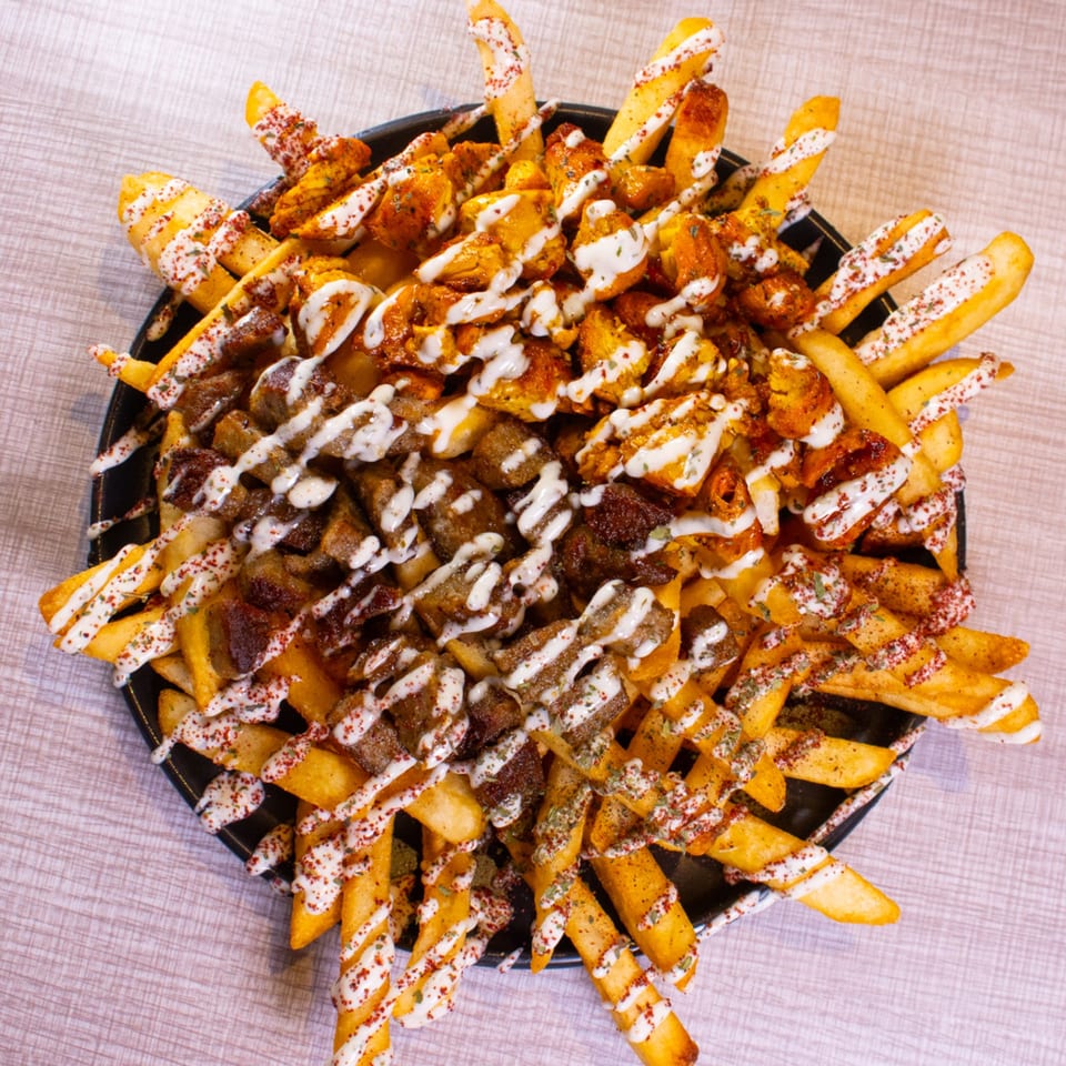 A black plate piled high with golden fries, chicken, beef, white sauce, and red spice.