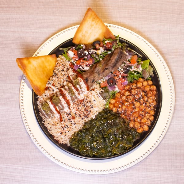 A colorful platter with rice, meat, salad, chickpeas, and greens, garnished with pita bread.