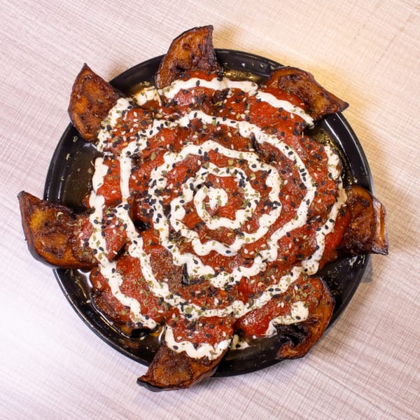 A black plate of fried plantains topped with red sauce, white drizzle, sesame seeds, and herbs.